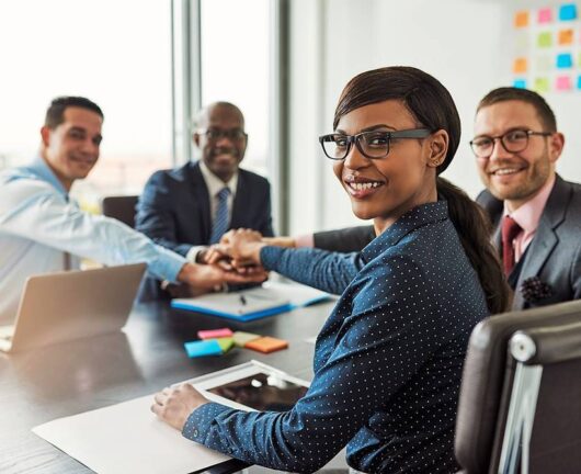 Successful African American team leader turning to smile at the camera as her multiracial team of executives links hands across the table